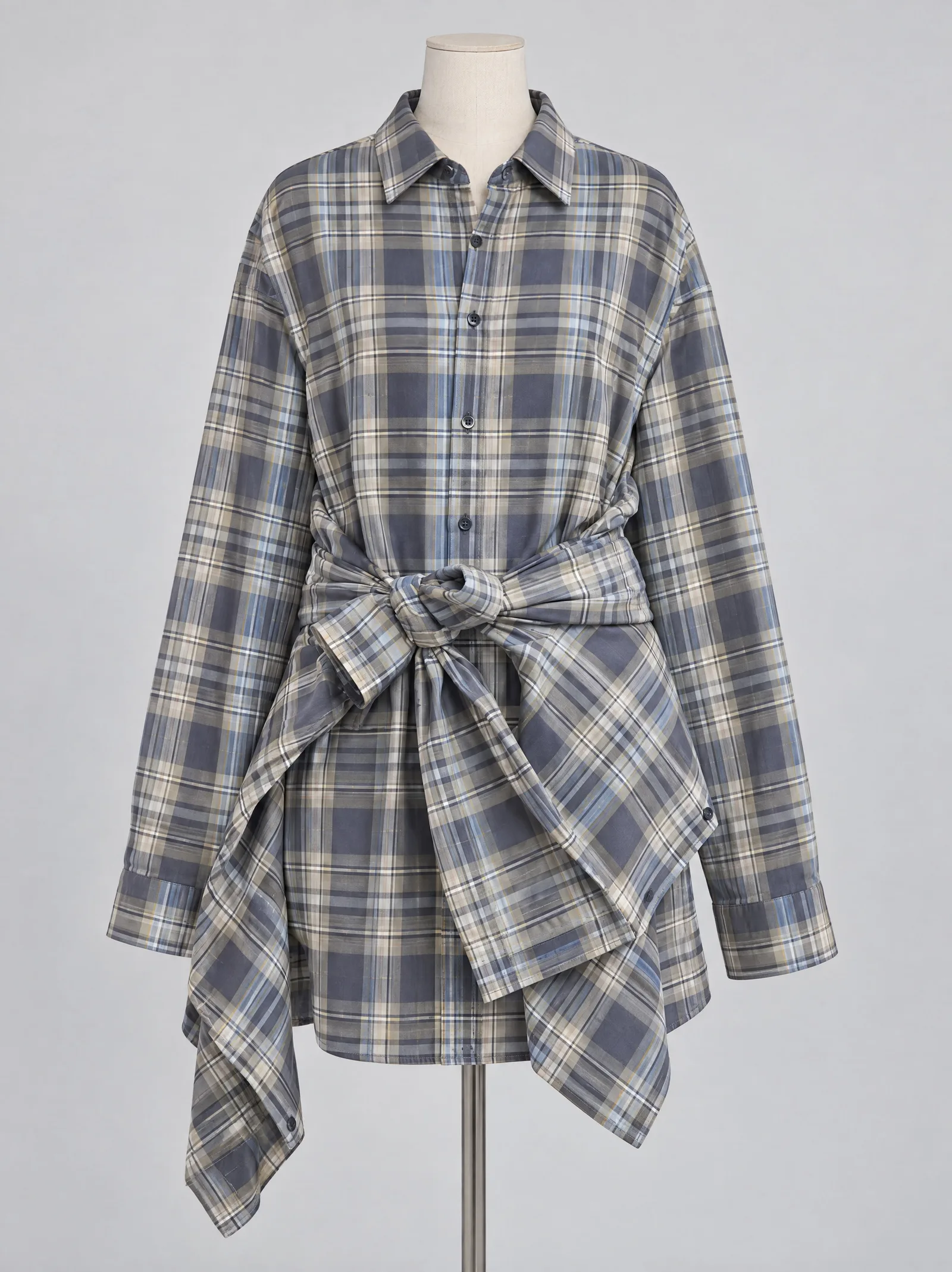 The same plaid shirt dress on a headless cream dressmaker's bust against a plain grey wall, a second matching shirt knotted at the waist