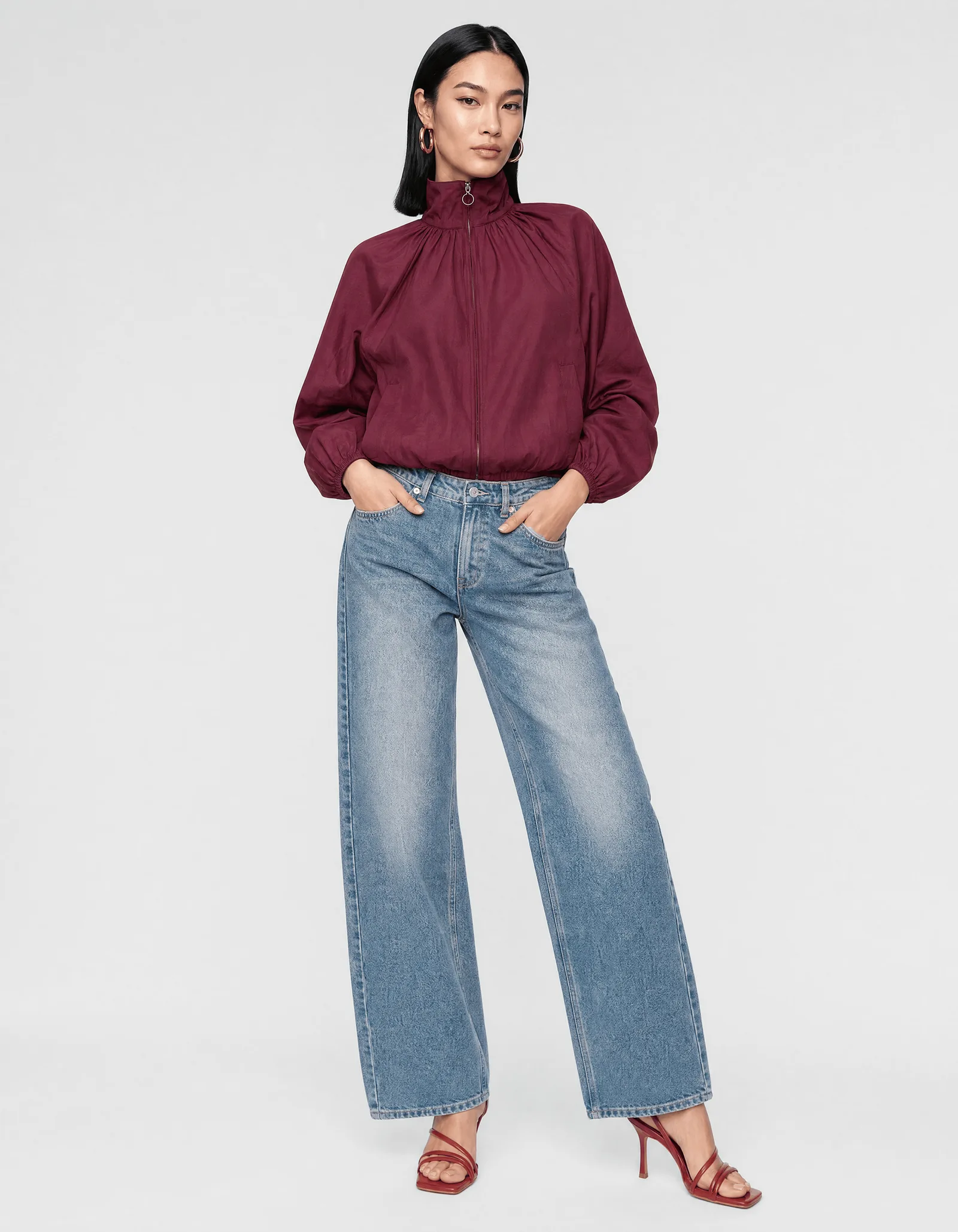 A model wearing the burgundy zip jacket with the light wash jeans, shot full length on a plain studio backdrop
